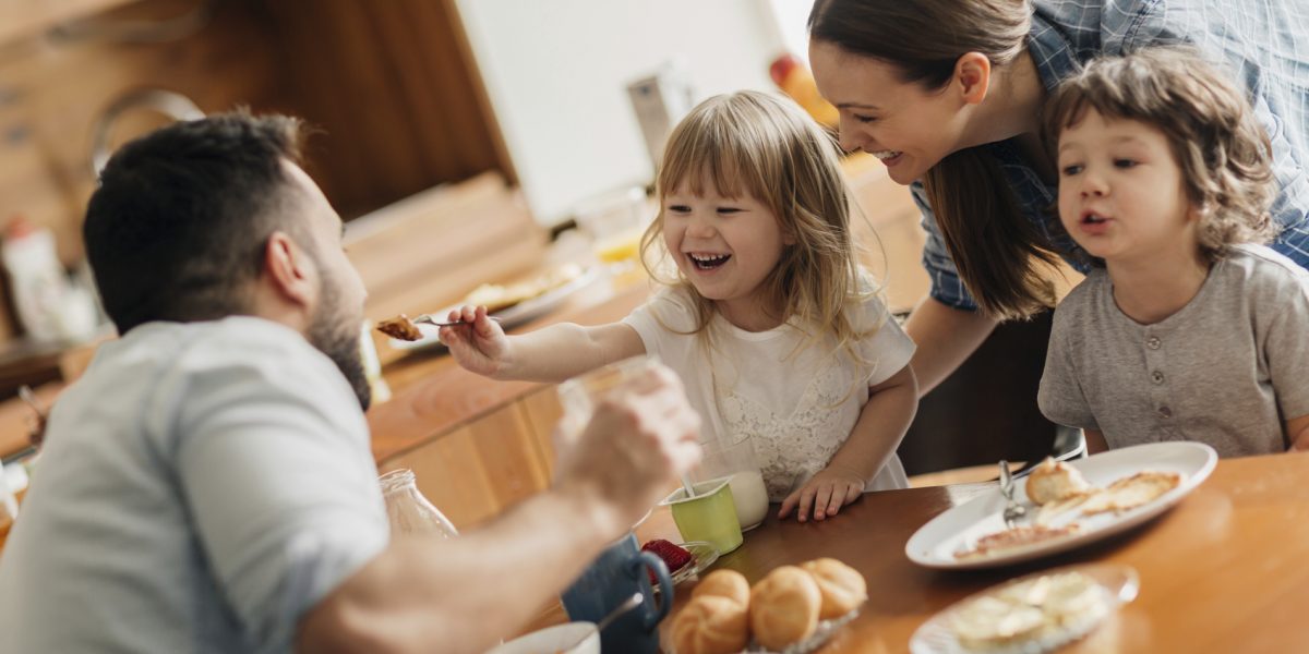 Picture of a young family enjoying their breakfast together. The children are all still in their pyjamas but the parents are dressed. On the breakfast table are pancakes, milk, cornflakes, croissants and strawberries. The daughter is playfully feeding her father and the mother and son are looking at them. In the background is the kitchen with wooden details.